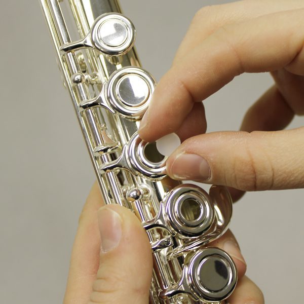 Close-up of hands playing a silver flute fitted with Jupiter Open-Hole Flute Plugs, fingers pressing down on its round keys against a neutral background.