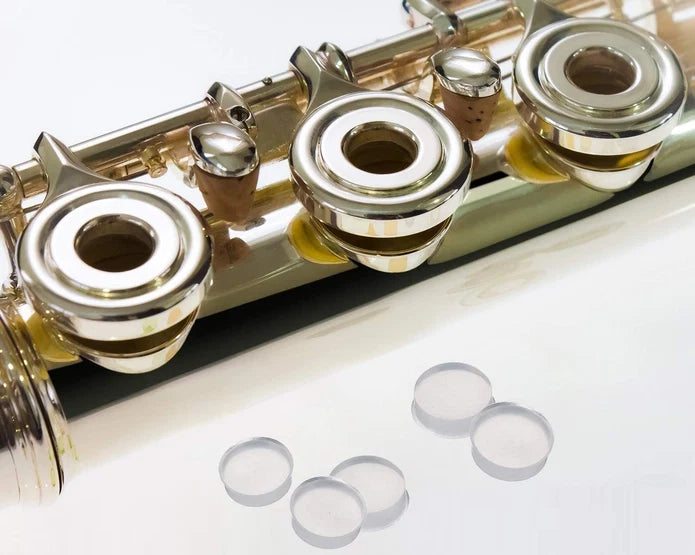 Close-up of a silver flute’s keys with several small, clear, circular key pads—Jupiter Open-Hole Flute Plugs—arranged on a white surface nearby, highlighting useful flute accessories like Jupiter Open-Hole Flute Plugs for customizing your instrument.