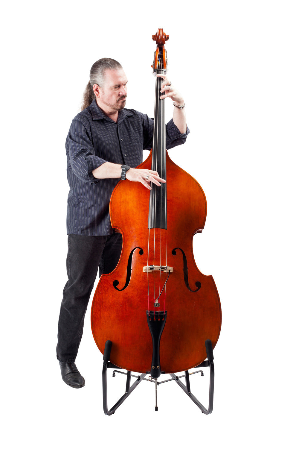 A man with long hair pulled back plays a large double bass on a stand. He wears a dark shirt and black pants, standing against a white background.