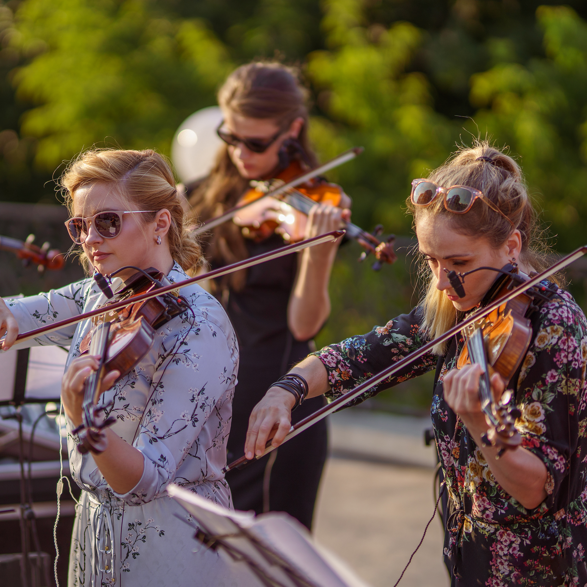 Three women wearing sunglasses play violins outdoors on a sunny day. They are focused on their music, with green foliage blurred in the background.