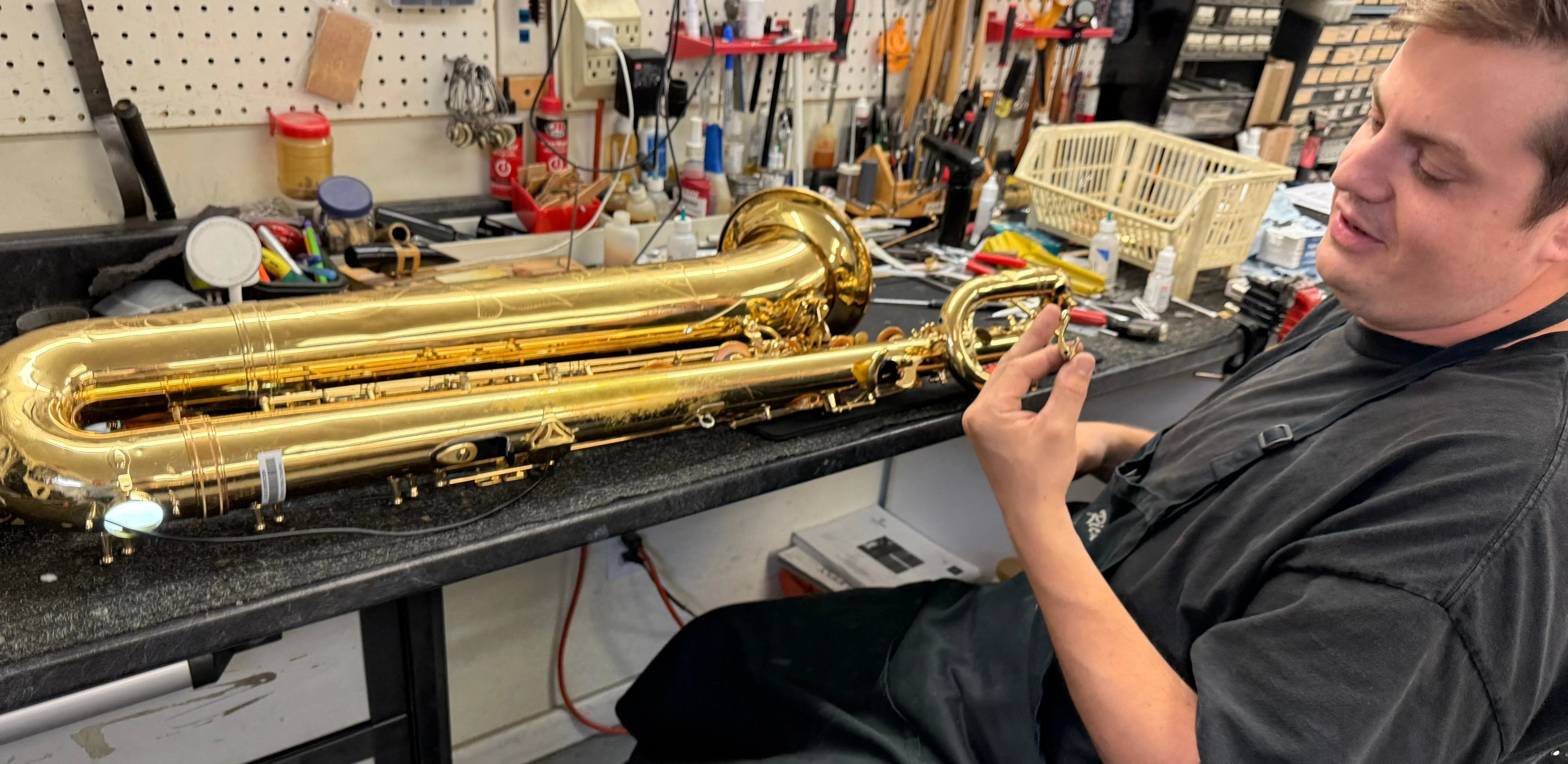 A person sits at a workbench, inspecting a large brass saxophone surrounded by tools and repair equipment in a workshop setting.