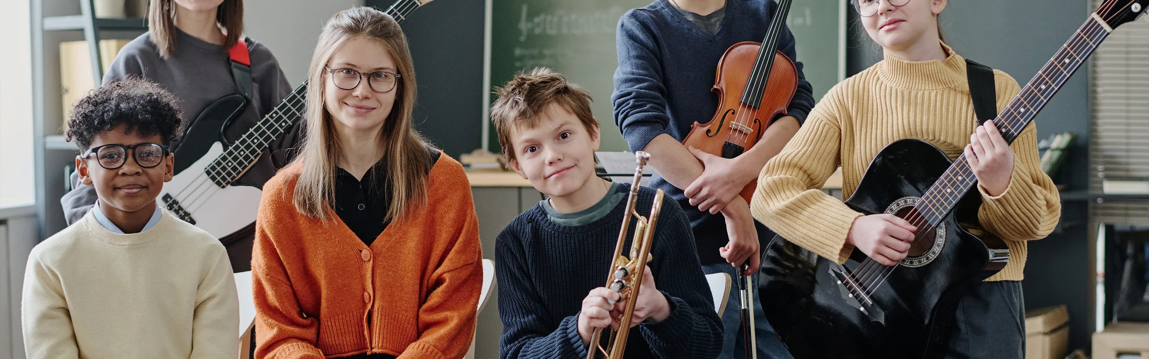 A group of children in a classroom, each holding a musical instrument, including a guitar, violin, trumpet, and bass guitar, smiling and posing for the photo. A chalkboard is visible in the background.