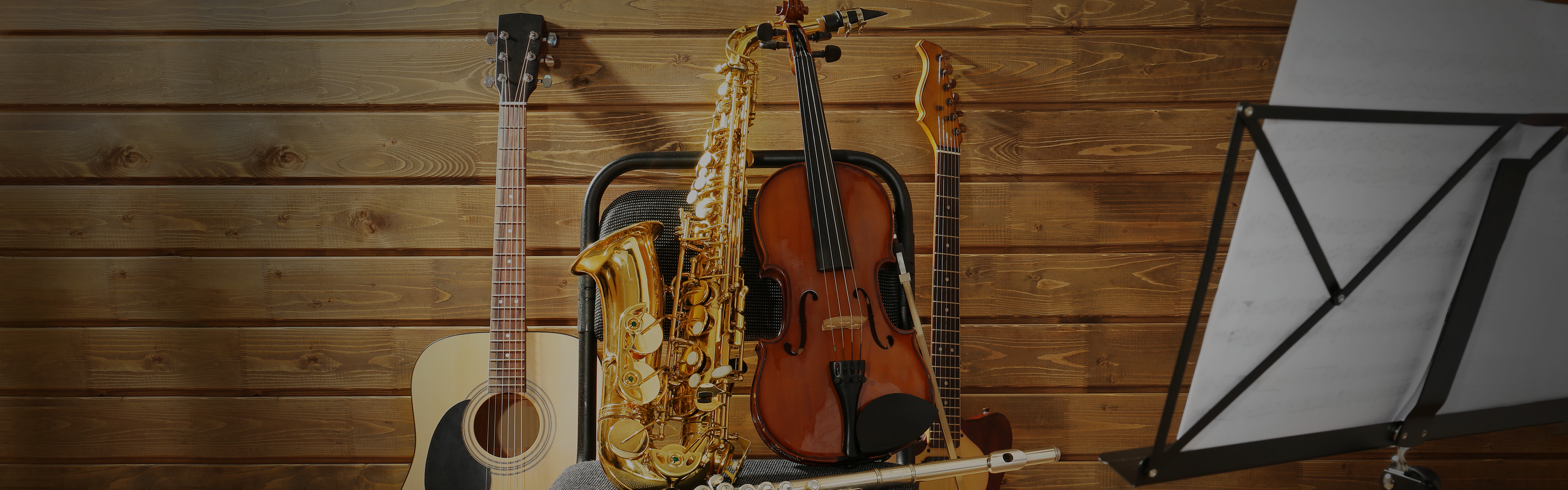A guitar, saxophone, violin, and flute are arranged in front of a wooden wall, next to a music stand holding sheet music.