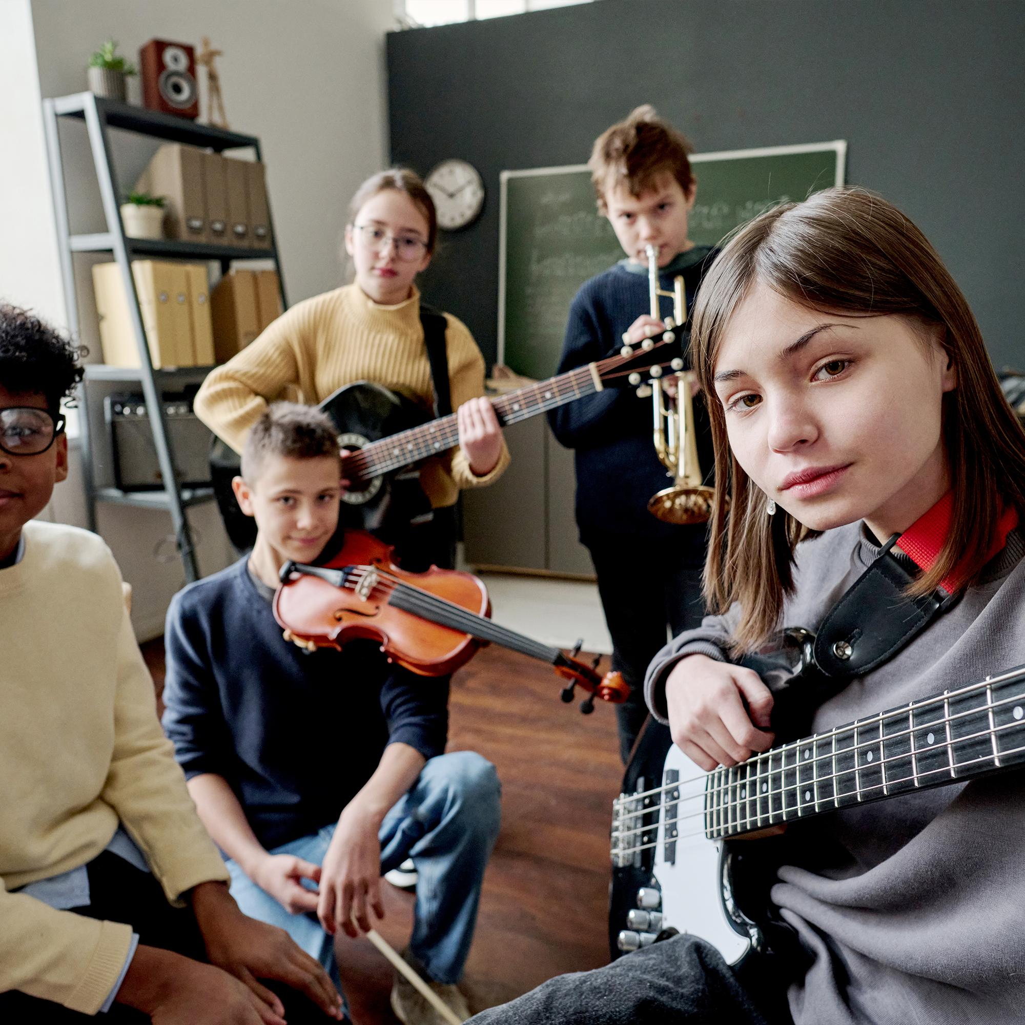 A group of five students in a classroom hold musical instruments—guitar, violin, bass, and trumpet—while posing for the camera. Shelves and a chalkboard are visible in the background.