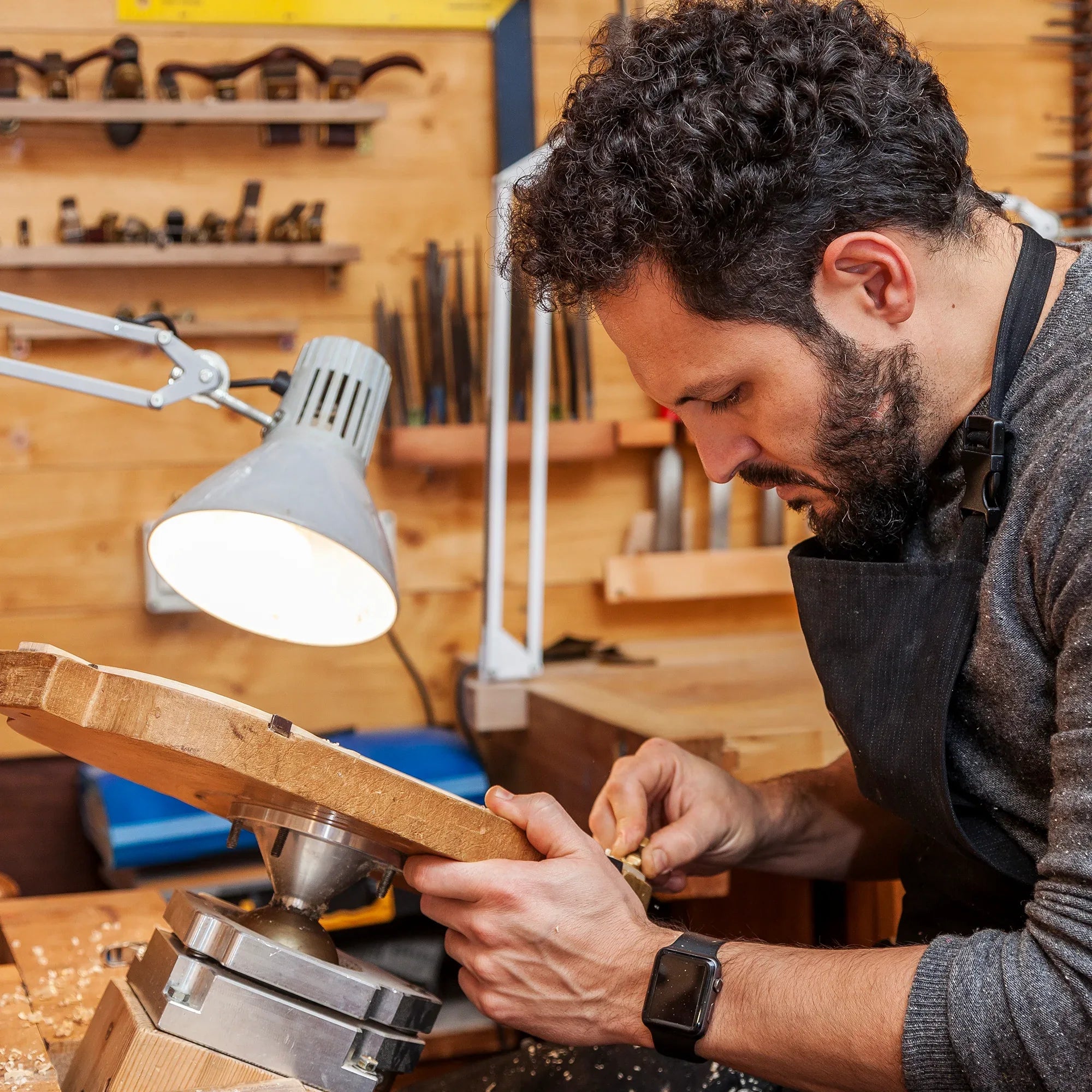 A man wearing a black apron and smartwatch carefully carves wood in a workshop, using a hand tool. A desk lamp and various woodworking tools are visible in the background.