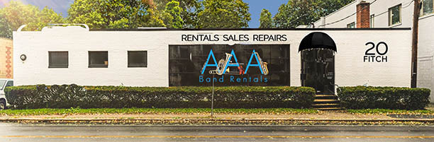 A white building with large windows labeled RENTALS SALES REPAIRS and AAA Band Rentals on the front. The address 20 Fitch is displayed on the right. There are trees and a wet street in front of the building.
