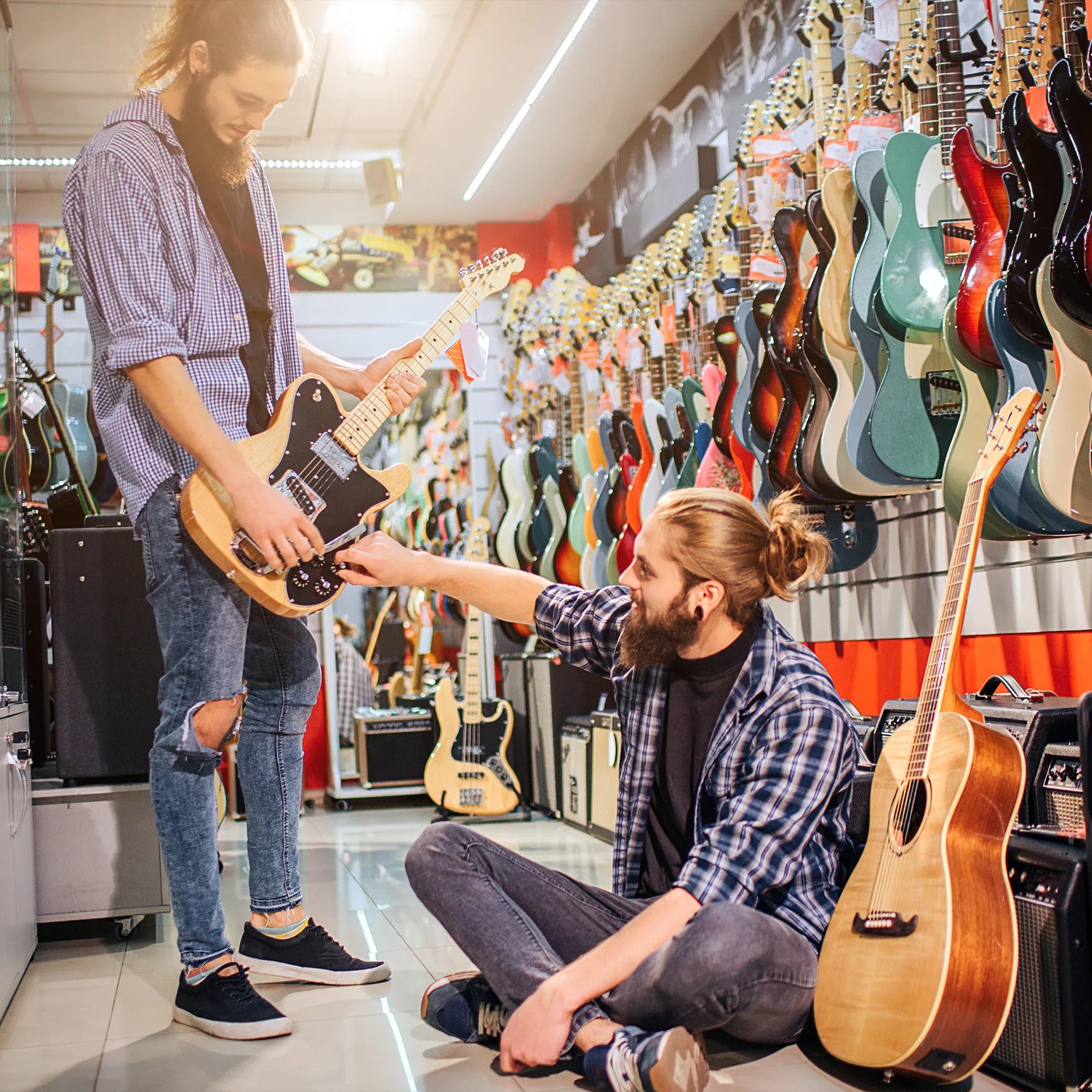 Two young men with long hair and beards interact in a guitar shop. One stands holding an electric guitar, while the other sits on the floor, reaching out to adjust the guitars settings. Colorful guitars hang on the walls around them.