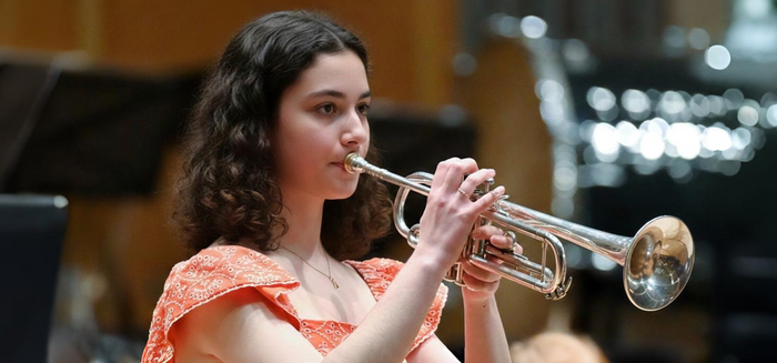 A young woman with curly brown hair, wearing an orange dress, plays a silver trumpet. She is focused on her performance, with a blurred background suggesting a concert or recital setting.