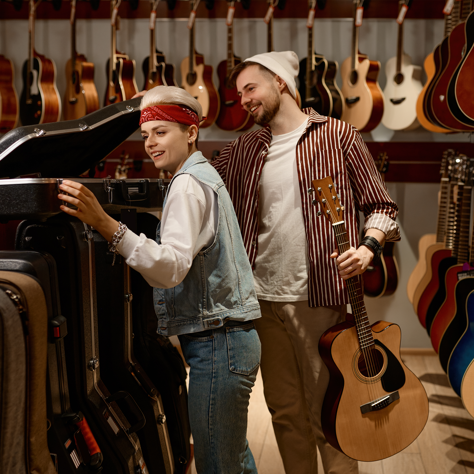 Two people in a guitar shop, one holding a guitar and smiling while the other opens a guitar case. Various acoustic guitars are displayed on the wall in the background.