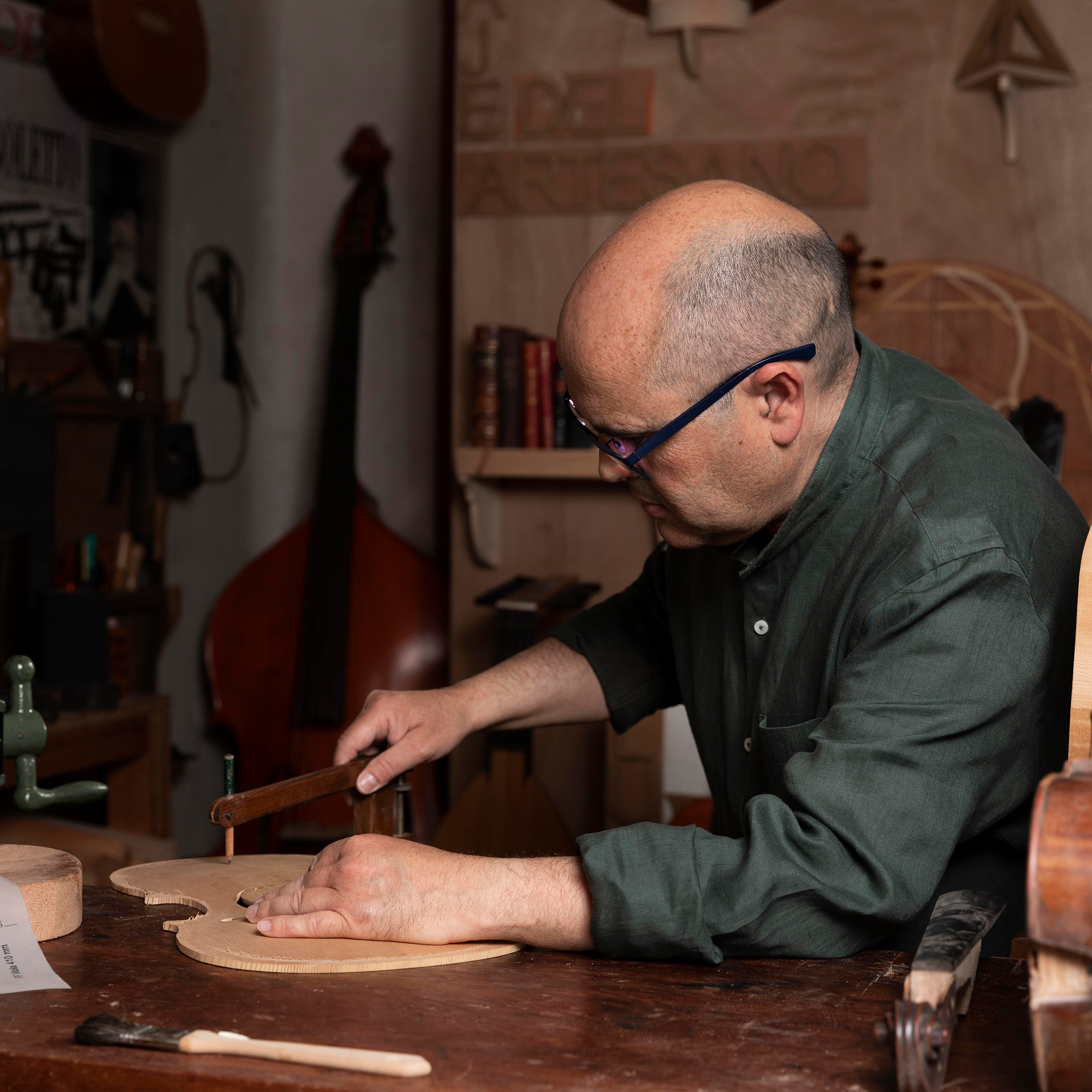 A man with glasses and a green shirt carefully carves a wooden violin body in a workshop filled with musical instruments and woodworking tools.