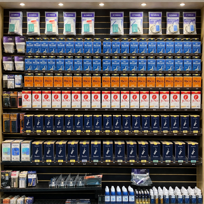A store display shows neatly organized rows of woodwind instrument reeds, mouthpieces, and accessories in various brands and colors, including Yamaha, Rico, Royal, and Juno, on shelves with bright lighting above.