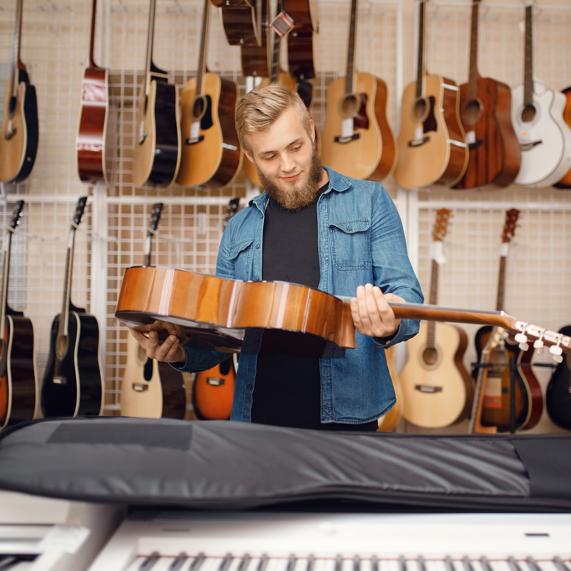 A man with a beard examines an acoustic guitar in a music store, surrounded by guitars hanging on the wall and a keyboard in the foreground.
