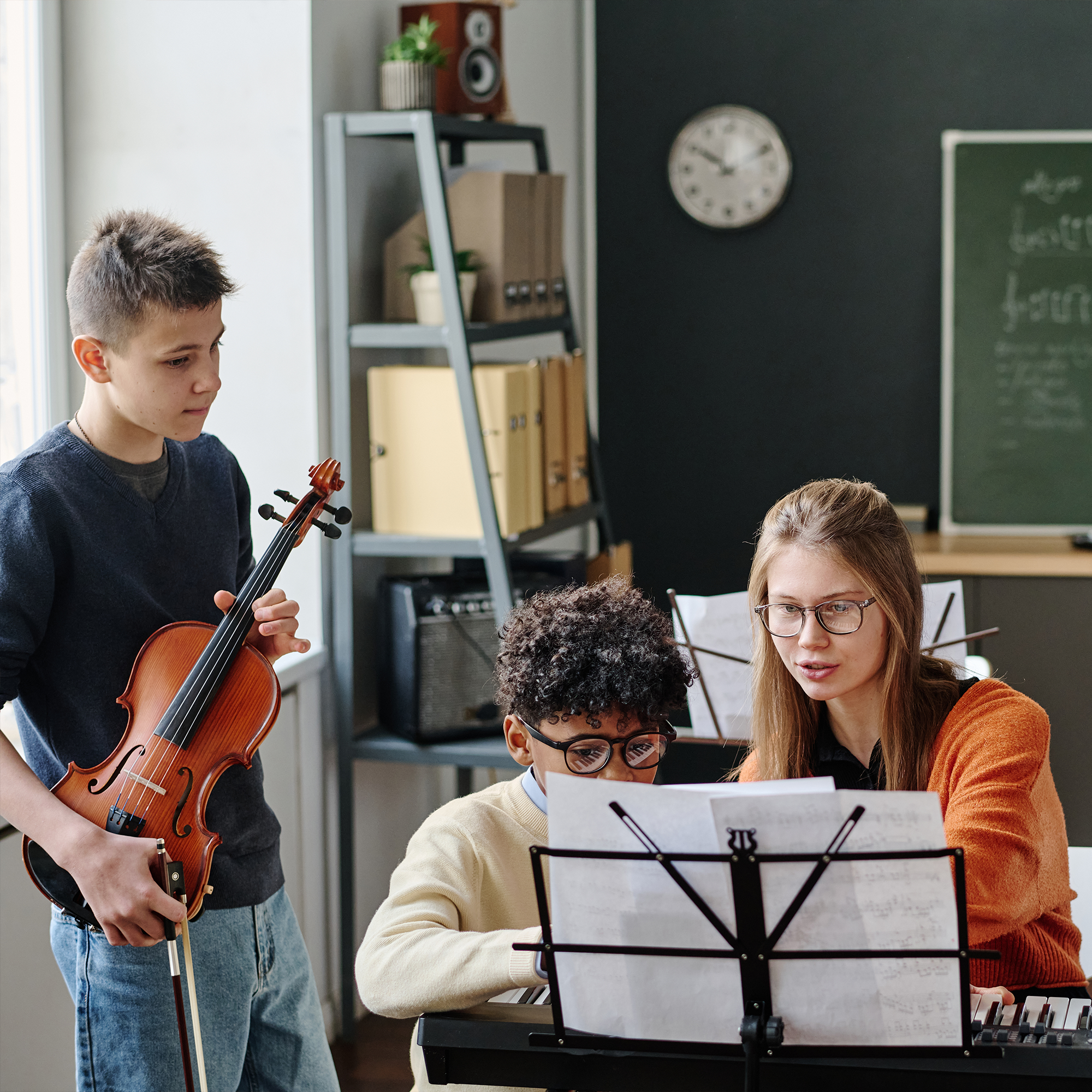 A teacher helps two students with sheet music at a keyboard; one student plays the keyboard while the other holds a violin. Shelves, a chalkboard, and a clock are visible in the background.
