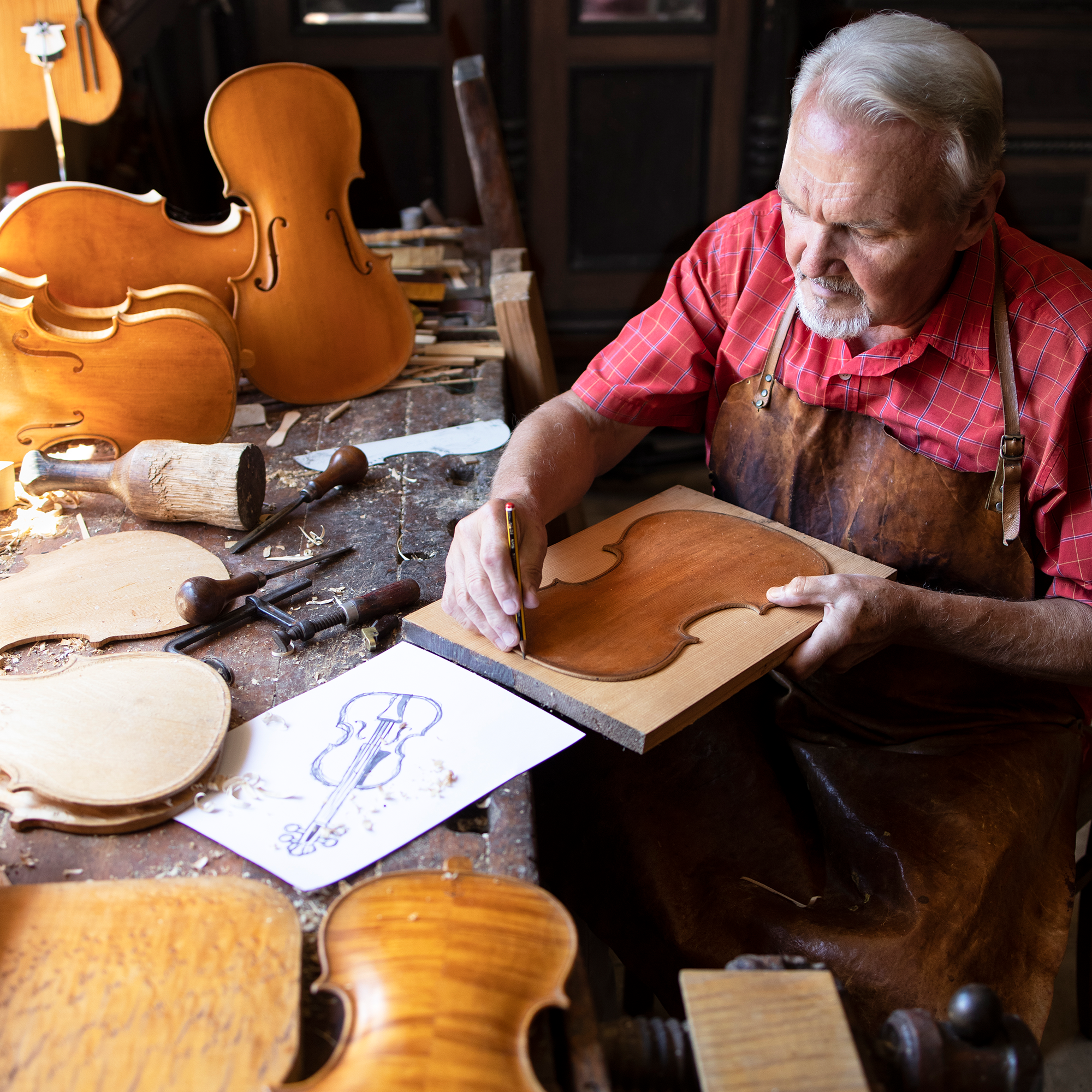 An elderly man in a red plaid shirt and apron works on a violin piece at a cluttered workbench, surrounded by violins, tools, wood, and a sketch of a violin.