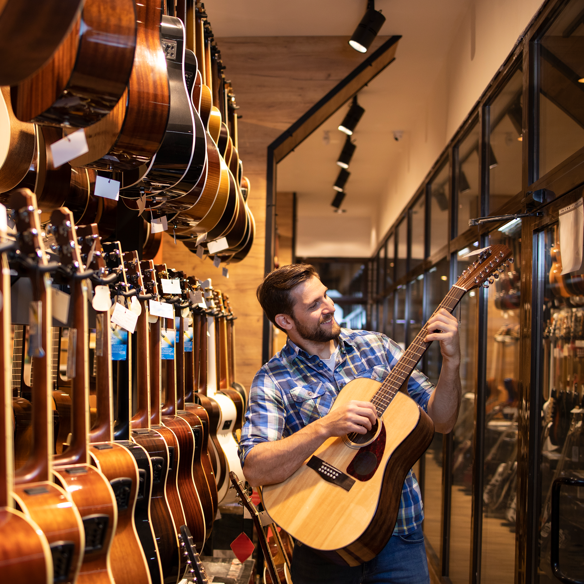A man in a plaid shirt smiles while playing an acoustic guitar in a music store, surrounded by rows of hanging guitars on both sides.