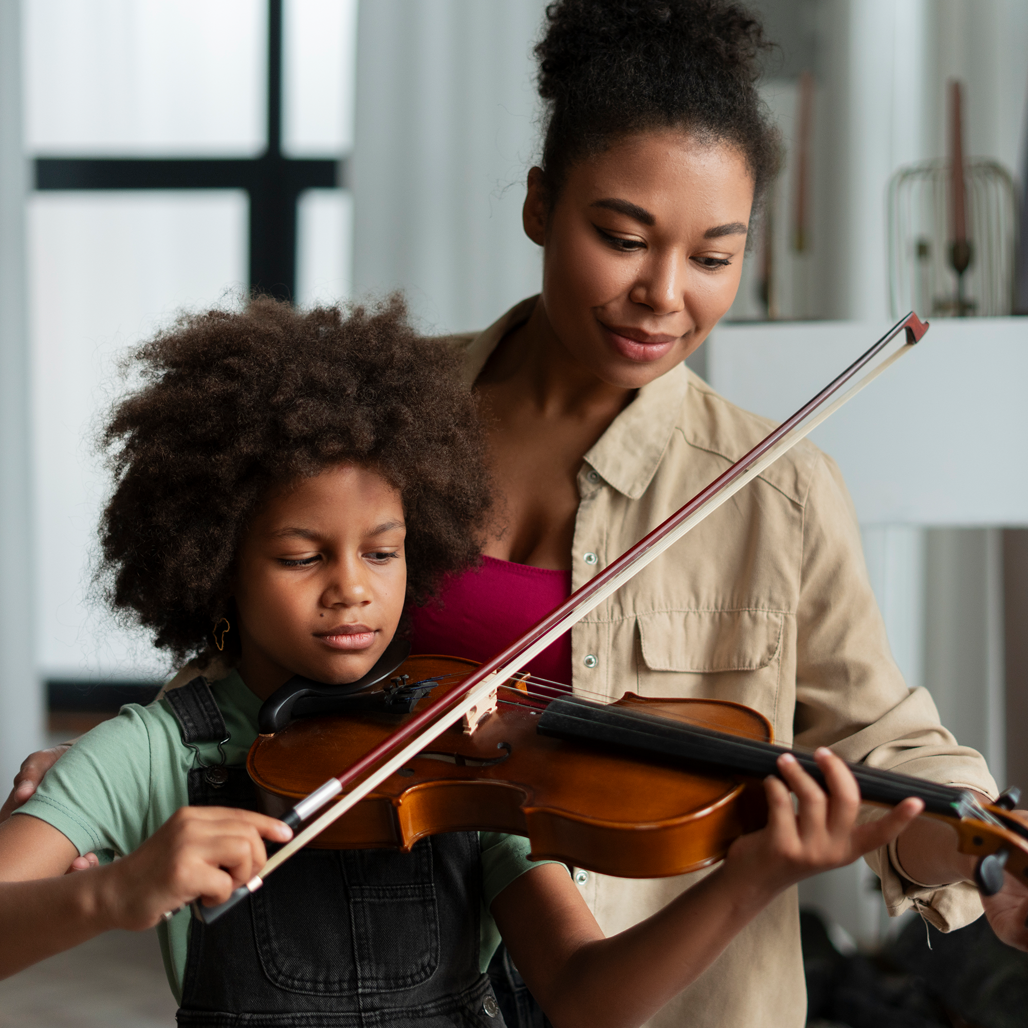 A woman helps a young girl play the violin, guiding her hands while the girl focuses on the instrument. They are indoors, and natural light comes through a window in the background.