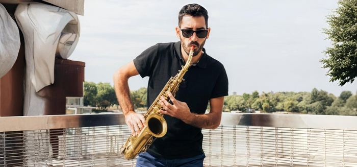 A man wearing sunglasses and a black polo shirt plays a saxophone outdoors, standing by a metal railing with trees and a cloudy sky in the background.