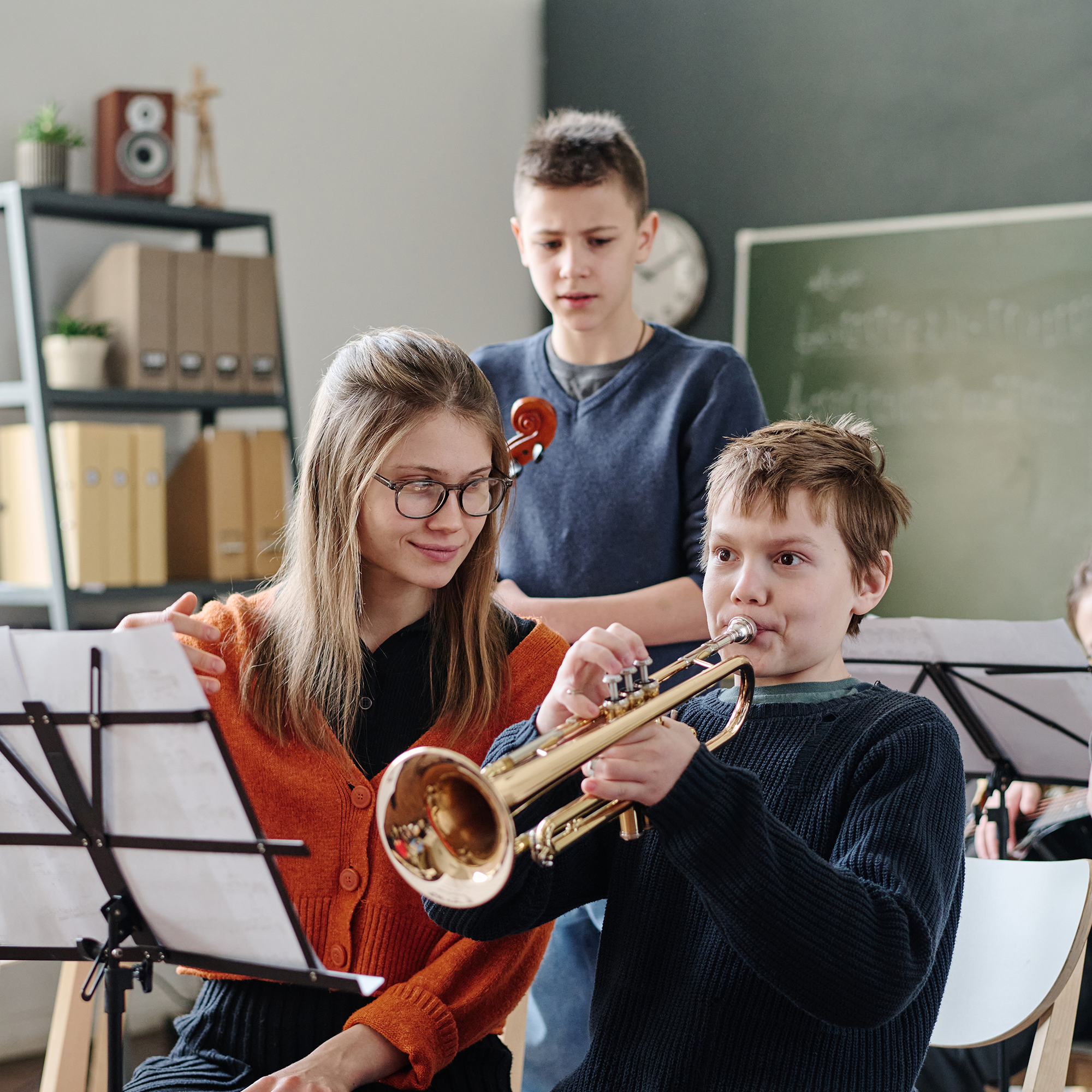 Three children in a classroom practice music; one plays a trumpet, another holds sheet music, and a third stands behind with a violin. Shelves, a clock, and a chalkboard are visible in the background.